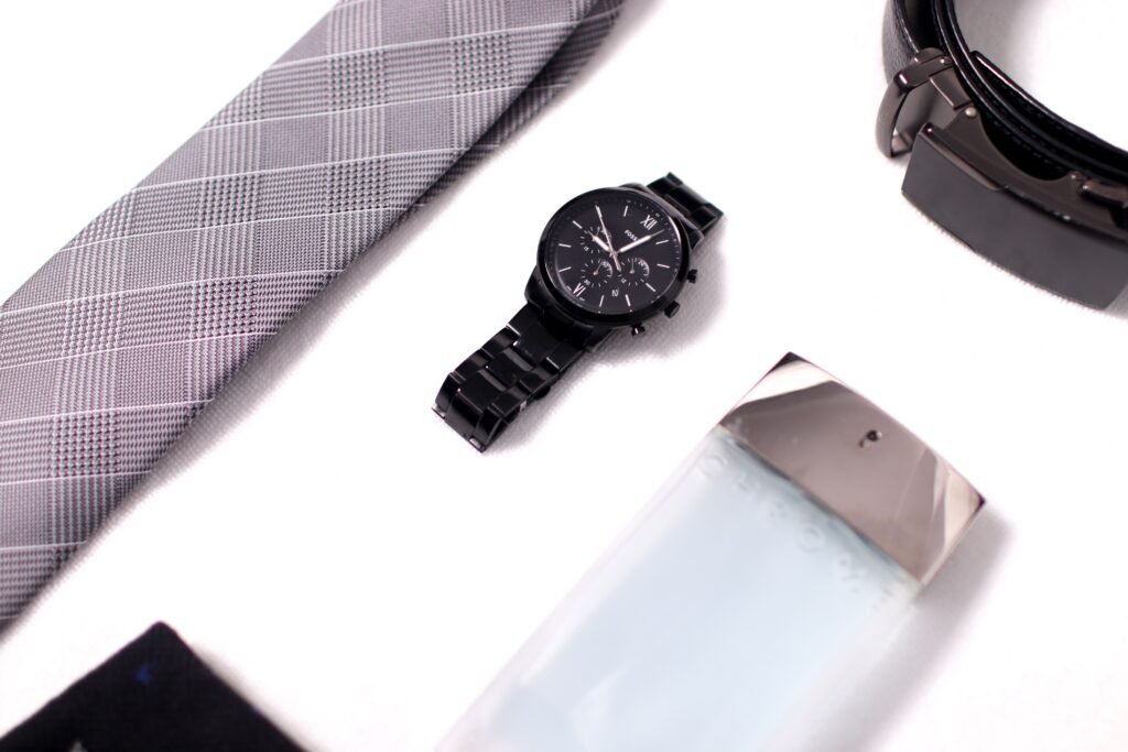 Flat lay of men's fashion items including a watch, tie, belt, and cologne.